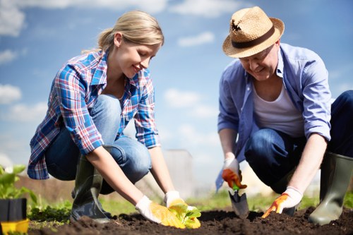 Two individuals, a young woman and an older woman, are engaging in gardening activities in a well-maintained outdoor space, likely part of a residential garden in Golders Green. The young woman, dressed casually in a blue and red checkered shirt and blue jeans, is kneeling on the soil, planting a small flowering plant with yellow blossoms. The older woman, wearing a straw hat, a light blue shirt, and dark trousers, is also kneeling and using gardening tools, possibly a trowel, to help with planting. The garden features a neat expanse of dark, fertile soil bordered by a paved path, with green grass and hedges visible in the background. Bright sunlight and a partly cloudy sky illuminate the scene, highlighting the fresh and lively garden environment. The setting suggests careful landscaping and planting maintenance typical of professional gardening services in the Golders Green area, emphasizing the natural tones of earth, green foliage, and flowering plants, while capturing an active outdoor gardening scene.