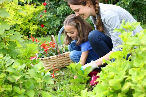 A woman and a young girl are gardening together in a lush outdoor space, with the woman crouched down beside the girl who is holding a wicker basket. They are surrounded by vibrant green foliage, including flowering plants and shrubs, with some red blossoms visible. The garden features a mix of well-maintained grass, soil beds, and flowering borders, all under natural daylight. In the background, there's a hedge or shrubbery creating a private outdoor environment, indicating a landscaped yard typical of residential gardens in Golders Green. The scene conveys a moment of outdoor activity and garden maintenance, with a focus on planting and nurturing garden elements, aligning with gardening and landscaping services in the area.