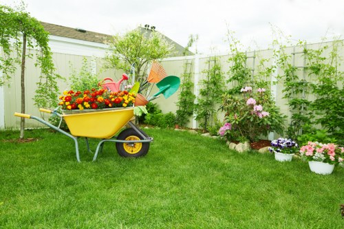 A well-maintained front garden features a vibrant green lawn bordered by a white fence, with various flowering plants and shrubs along the edges. In the foreground, a yellow wheelbarrow filled with colourful garden flowers and gardening tools such as a watering can and garden fork is positioned on the grass. To the right, a selection of potted plants with pink, purple, and white blooms adds visual interest and colour contrast. Behind the flower beds, a small garden hedge and a shrub with sparse branches are visible, indicating active gardening care. The environment appears bright and clear, suggesting a mild weather day ideal for outdoor gardening activities. The scene is typical of a residential landscape in the Golders Green area, showcasing a neat and inviting outdoor space suitable for various gardening and landscaping services offered by Landscapers Golders Green, including lawn maintenance, planting, and garden beautification in the local community.