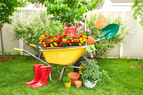 A garden scene in a residential outdoor space features a central yellow wheelbarrow filled with vibrant red, orange, and yellow flowers, positioned on a well-maintained green lawn. Surrounding it are gardening tools and accessories, including a red watering can, a pair of red rubber boots, and several small flower pots in shades of terracotta and yellow, placed on the ground. To the right of the wheelbarrow, there is a potted plant with lush green foliage, along with a small garden trowel and a green watering can, indicating active gardening. Large trees and bushy shrubs border the garden in the background, providing shade and privacy, with their leaves casting dappled light on the scene. A white fence encloses the space, enhancing the sense of an organized, cultivated outdoor area suitable for gardening services in Golders Green and nearby NW postcode areas. The overall setting suggests a neat, well-kept garden during daylight hours with natural, soft sunlight highlighting the vivid colours and textures of the plants and tools, reflecting typical gardening and landscaping maintenance tasks.