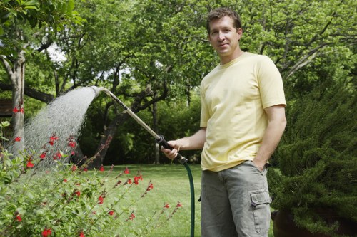 A man standing in a well-maintained garden in Golders Green, London, watering plants with a garden hose. The garden features a lush, green lawn, vibrant flower beds with red flowers, neatly trimmed hedges, and mature trees in the background. The scene is outdoors on a bright, partly cloudy day, with natural daylight illuminating the various plants and garden elements. The man is casually dressed in a light yellow T-shirt and grey cargo trousers, displaying a relaxed posture as he cares for his garden. The landscaped outdoor space emphasizes tidy borders, healthy foliage, and a peaceful environment, reflecting professional gardening and landscaping standards typical of services offered by Landscapers Golders Green. This outdoor setting is an example of meticulous garden management, showcasing the potential of regular lawn care, planting, and garden maintenance services available in the London NW11 area.