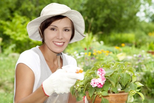 The image shows a woman in a white hat and gardening gloves in a lush garden environment, tending to a potted flowering plant with pink blossoms. She has short brown hair and a smile, wearing a white short-sleeved top. The background includes a variety of green foliage, flowering plants, and shrubs, with sunlight illuminating the scene, indicating a bright and mild weather day. The garden is organized with a mix of flower beds, lawn areas, and natural greenery, typical of residential outdoor spaces that benefit from professional gardening and landscaping services. This setting reflects the natural beauty and outdoor maintenance focus relevant to the services offered by Landscapers Golders Green in the NW postcode area, especially around Golders Green in London, supporting lawn care, planting, and garden upkeep activities.