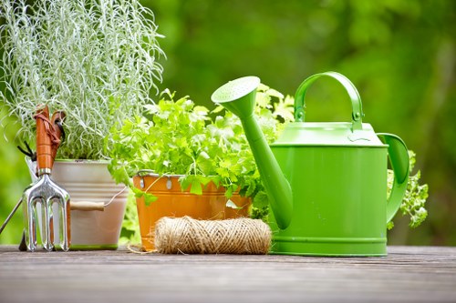 A close-up of a wooden garden table displays various gardening tools and plants, including a green watering can with a long spout, a small terracotta pot with leafy green herbs, and a white pot containing a tall, silvery-grey plant. Additionally, there is a gardening fork with a wooden handle and metal tines resting against the pots, along with a neatly coiled piece of jute twine. The background features a lush, out-of-focus garden with vibrant green foliage and natural daylight, suggesting a well-maintained outdoor space typical of a residential garden in Golders Green. This arrangement highlights outdoor gardening activities and tools that landscapers in the area, like Landscapers Golders Green, use when creating flower beds and maintaining garden beds, lawns, and general landscape features.