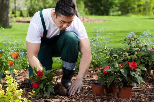 A gardener wearing a white t-shirt and green overalls is kneeling on a mulch bed in a lush garden, actively planting or tending to red and yellow flowering plants in terracotta pots and directly in the soil. Surrounding the scene are vibrant green grass, shrubbery, and small blooming flowers, with a background of mature trees and a well-maintained lawn, indicative of a landscaped outdoor space in the Golders Green area. The natural daylight highlights the rich textures of the mulch, foliage, and flowers, reflecting careful garden maintenance and planting work typical of professional landscaping services offered by Landscapers Golders Green.