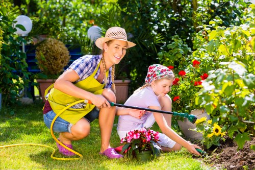 A woman and a young girl are gardening together in a lush backyard. The woman, wearing a straw sunhat, yellow apron, checkered shirt, and denim shorts, is smiling while holding a garden hose, watering a flower bed. The young girl, dressed in a purple T-shirt, light-colored pants, and a floral hat, is kneeling on the grass, tending to a cluster of pink and yellow flowering plants. The garden features a well-maintained lawn with vibrant green grass, bordered by flowering plants and bushes, including red and yellow blooms. In the background, there are tall green hedges, small trees, and garden decorations such as white spherical lights hanging from a trellis, creating a cheerful and inviting outdoor space typical of a private garden in Golders Green. The weather appears sunny with dappled sunlight filtering through the foliage, highlighting the natural tones of the plants and the vibrant colors of the flowers, demonstrating a well-kept garden suitable for outdoor activities and gardening services provided by Landscapers Golders Green.