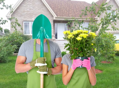 In a residential garden, two individuals are preparing for gardening activities; one wears a teal gardening helmet, a grey t-shirt, an eye-catching green apron, and yellow gloves, holding a garden shovel upright, while the other, dressed in a grey top with pink gloves, is partially obscured behind a large pink pot filled with bright yellow flowers, possibly chrysanthemums, with lush green foliage. The garden features a well-maintained lawn with dense, healthy grass, bordered by a variety of shrubs and small trees, and a modern brick house with white-framed windows and a sloped roof in the background. The scene suggests outdoor gardening work in a suburban setting, with natural daylight illuminating the landscape, highlighting the vibrant colours of the plants and the greenery surrounding the area. This outdoor space is indicative of professional gardening or landscaping services, such as those offered by Landscapers Golders Green in the Golders Green area, promoting outdoor beautification and garden maintenance.
