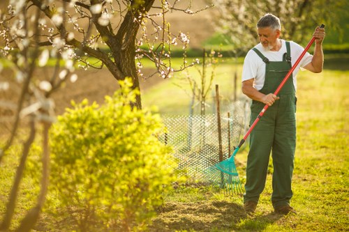 A middle-aged man wearing green gardening overalls and a white t-shirt is standing in a well-maintained backyard garden in Golders Green, trimming a small shrub with a rake, with a background of blooming trees with white flowers and a lush green lawn. The garden features a mix of grass, soil, and garden beds bordered by low fencing, with various plants and flowering bushes. The sunlight creates a warm and inviting atmosphere, highlighting the natural tones of the foliage and the textured paving of a nearby patio area. This outdoor space showcases typical landscaping elements suitable for garden maintenance and enhancement by professional gardening services in the Golders Green area, emphasizing a tidy and aesthetically pleasing garden environment.